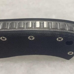 May include: Close-up of a black folding knife handle with a textured surface and silver screws. The handle has a silver metal inlay with a patterned design.