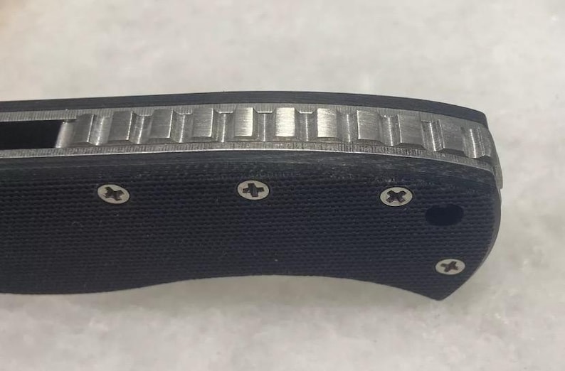 May include: Close-up of a black folding knife handle with a textured surface and silver screws. The handle has a silver metal inlay with a patterned design.
