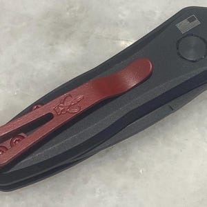 May include: A black pocket knife with a red pocket clip. The knife has a small American flag design on the handle.