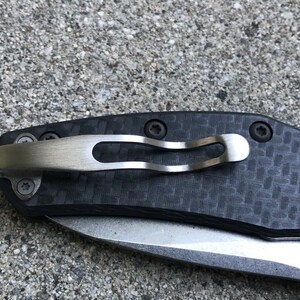 May include: A close-up of a black pocket knife with a carbon fiber handle and a silver pocket clip. The knife has a partially open blade and the text "L.T.R.O.C.K.  E.A.R. PRIMITIVE  MADE IN USA" is visible on the blade.