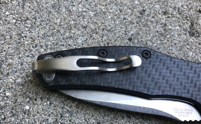May include: A close-up of a black pocket knife with a carbon fiber handle and a silver pocket clip. The knife has a partially open blade and the text "L.T.R.O.C.K.  E.A.R. PRIMITIVE  MADE IN USA" is visible on the blade.