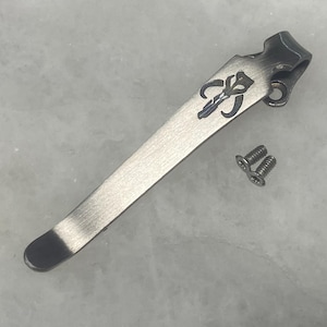 May include: A silver-toned pocket clip featuring a Mandalorian symbol and two small screws. The clip has a curved design with a dark finish on the end and the top. The Mandalorian symbol is in blue and black.