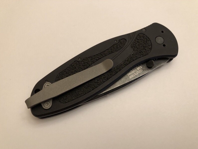 May include: A black folding knife with a textured handle and a pocket clip. The blade is partially open and the knife has the text "MISTRSTM" and "R2" printed on the side.