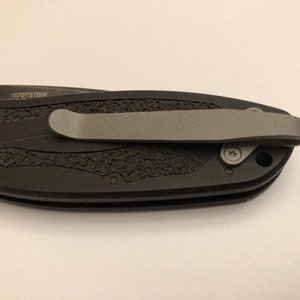 May include: Black pocket knife with a textured handle and a silver pocket clip. The knife has a small screw on the side and the text "1670TSTBW" is engraved on the handle.