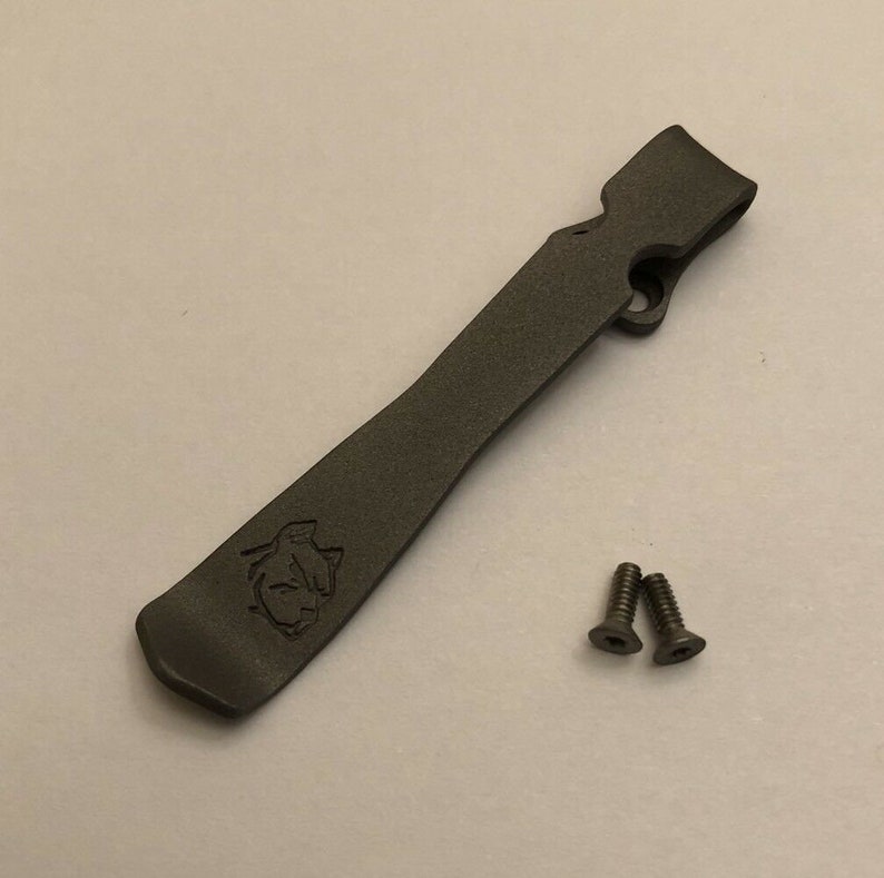Puede incluir: Un clip de bolsillo de metal gris oscuro con un logotipo y dos tornillos peque&ntilde;os.