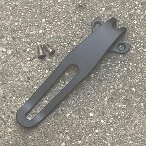 May include: A matte black metal pocket clip with a long, narrow central cutout and a flared mounting base featuring two circular screw holes, accompanied by two small silver-colored metal screws with flat heads for secure attachment.