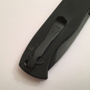 May include: Black pocket knife with a black pocket clip. The pocket clip has a logo on it.