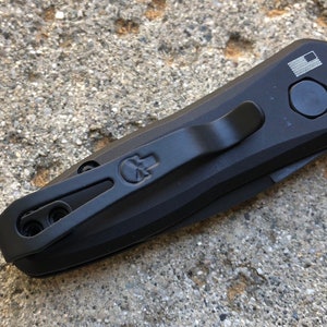 Puede incluir: Cuchillo plegable negro con un logotipo de calavera y un clip de bolsillo. El cuchillo tiene un mango negro y una hoja negra. El cuchillo tiene un botón en el lateral para abrir la hoja.