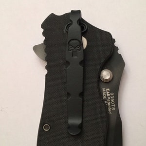 Pu&ograve; includere: Coltellino tascabile nero con un clip a forma di teschio e il testo "0350TS MADE Patented" sul lato.