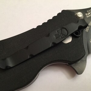 Pu&ograve; includere: Coltellino da tasca nero con clip nero e logo a forma di teschio. Il coltellino ha inciso sul lato il testo "0350TS KAI Patented MADE IN USA SER. 8887 Ken Onion Design S30V".