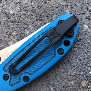 May include: A black pocket clip with a butterfly logo attached to a blue knife handle. The clip has a textured surface and is secured with screws.