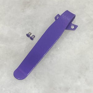 May include: A purple metal pocket clip with two small silver screws. The clip is a solid, curved piece with a rounded end and a loop at the top with two holes for mounting. The clip is designed to attach to a pocket or other item.