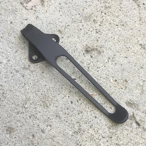 May include: A matte black metal door latch component featuring a long, slender arm with an elongated central cutout and a triangular mounting base with two circular screw holes for secure attachment to a door or frame surface.