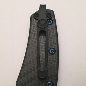 Puede incluir: Un clip de bolsillo negro con un dise&ntilde;o de calavera, unido a un mango de cuchillo de fibra de carbono negro. El clip tiene tornillos azules.