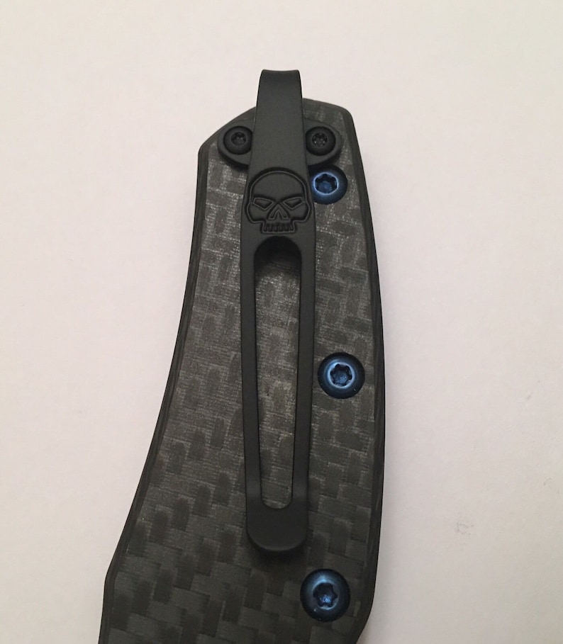 Puede incluir: Un clip de bolsillo negro con un dise&ntilde;o de calavera, unido a un mango de cuchillo de fibra de carbono negro. El clip tiene tornillos azules.