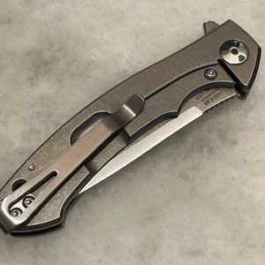 May include: A gray pocket knife with a partially serrated blade and a pocket clip. The knife has a black skull design on the handle and the text "0450 Kai USA" is engraved on the handle.