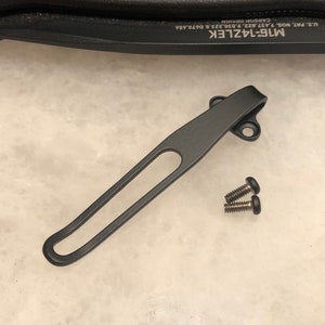 May include: A black metal pocket clip with a curved design and a slotted opening, accompanied by two small screws. The clip is designed for secure attachment and features a matte finish. The text "M16-14ZLEK" is visible.