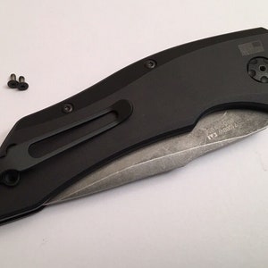 May include: Black folding knife with a partially open blade. The knife has a black handle with a pocket clip and a small American flag on the back. The blade is made of stainless steel and has a partially serrated edge. The knife is closed and has a small screw on the back.