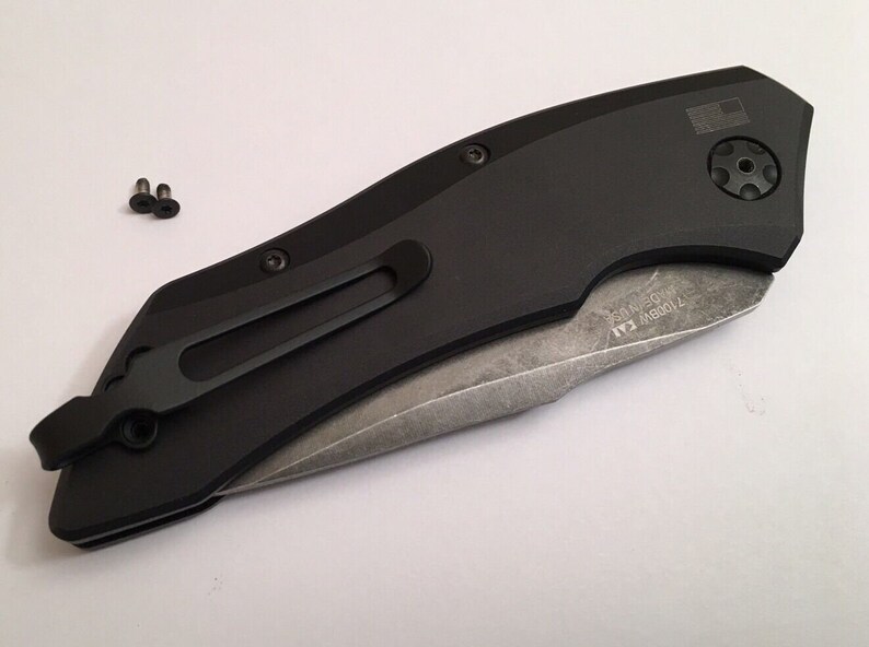 May include: Black folding knife with a partially open blade. The knife has a black handle with a pocket clip and a small American flag on the back. The blade is made of stainless steel and has a partially serrated edge. The knife is closed and has a small screw on the back.