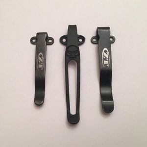 Puede incluir: Tres clips de bolsillo de metal negro con un logotipo ZT. El clip del medio tiene un dise&ntilde;o de calavera.