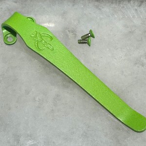 Puede incluir: Un clip de bolsillo de metal verde brillante con un diseño de libélula. Se incluyen dos pequeños tornillos plateados.