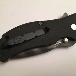 Pu&ograve; includere: Coltello pieghevole nero con un fermaglio da tasca nero e una vite del fermaglio da tasca argentata. Il coltello ha un'impugnatura nera e una lama argentata. La lama &egrave; parzialmente aperta. Il coltello ha il testo "03501S KAI brevettato MADE in" sul lato.