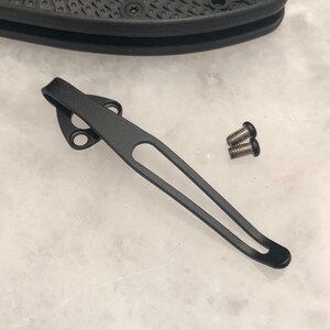 May include: Black metal pocket clip with two screws. The clip has a rounded end and a small hole in the middle.