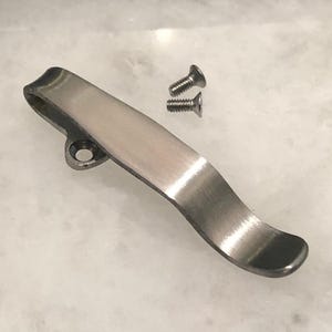 May include: A silver metal pocket clip with a curved design and a small hole at the top. Two small silver screws are included.