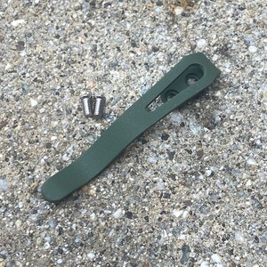 May include: A green metal pocket clip with two silver screws. The clip is designed to attach to a pocket or belt.