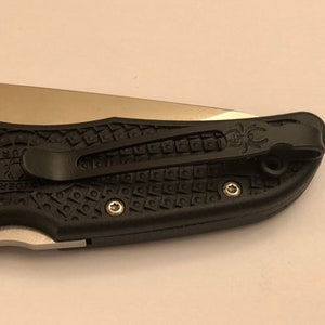 May include: A black pocket knife with a textured handle and a silver blade. The knife has a pocket clip with a spider logo.
