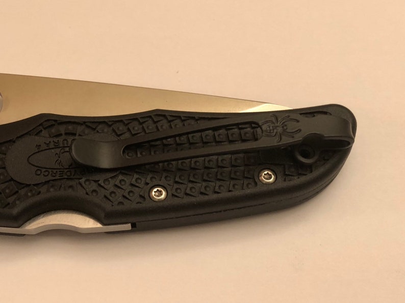 May include: A black pocket knife with a textured handle and a silver blade. The knife has a pocket clip with a spider logo.