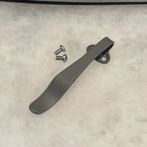 May include: A gray metal pocket clip with two silver screws. The clip is designed to attach to a pocket or belt.