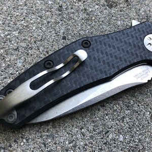May include: A black and silver pocket knife with a carbon fiber handle and a clip. The knife has a partially serrated blade and the text "MADE IN USA" is engraved on the blade.