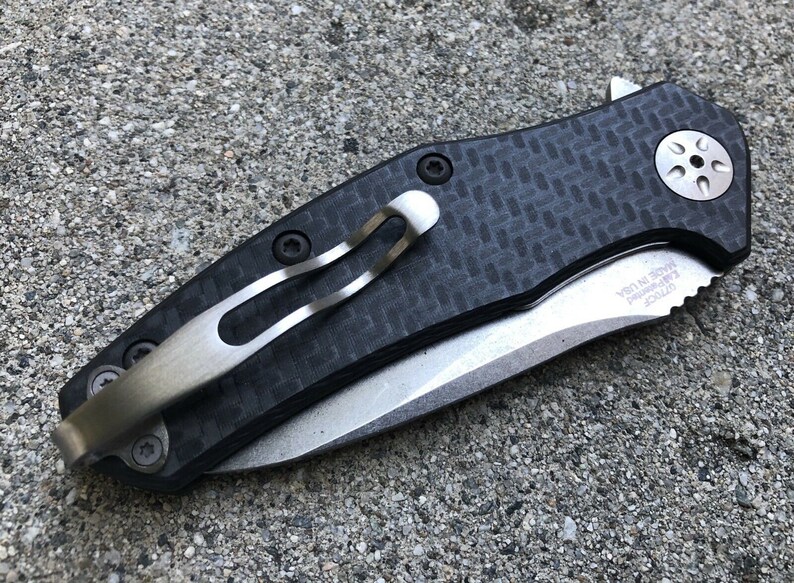 May include: A black and silver pocket knife with a carbon fiber handle and a clip. The knife has a partially serrated blade and the text "MADE IN USA" is engraved on the blade.