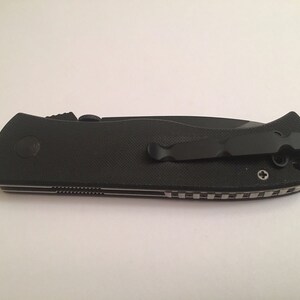May include: Black folding knife with a textured handle and a black pocket clip. The knife has a silver screw on the handle.