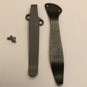 May include: Two black metal pocket clip parts for a knife. The larger part has the word "Kershaw" engraved on it. Two small silver screws are included.