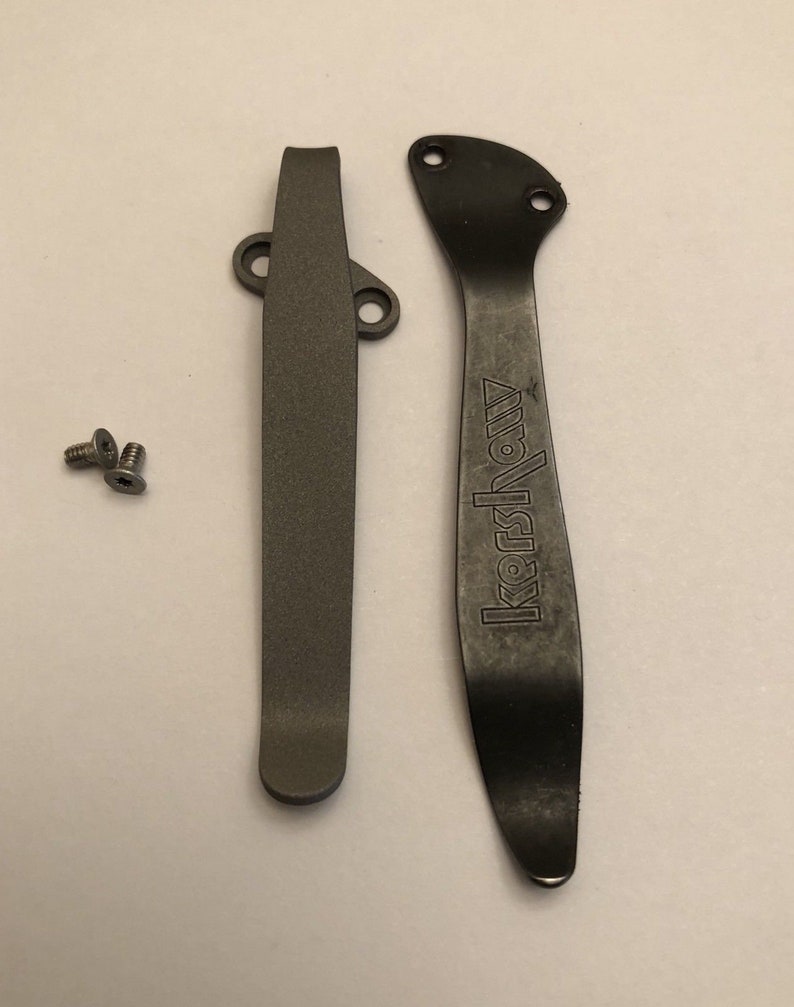 May include: Two black metal pocket clip parts for a knife. The larger part has the word "Kershaw" engraved on it. Two small silver screws are included.