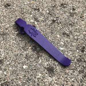 May include: A purple plastic keychain with a spider design. The keychain has a small loop at the top for attaching to keys.