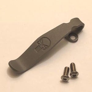 Puede incluir: Un llavero de metal gris con un diseño de calavera y dos tornillos.