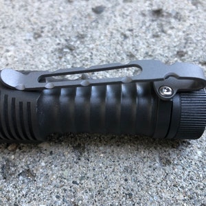 Puede incluir: Una linterna negra con un cuerpo texturizado y un clip de bolsillo plateado. El clip tiene un pequeño bucle en el extremo para sujetarlo a un cinturón o una bolsa.