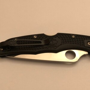 May include: A black folding knife with a silver blade and a black handle. The handle has a textured pattern and the Spyderco logo is visible on the clip.