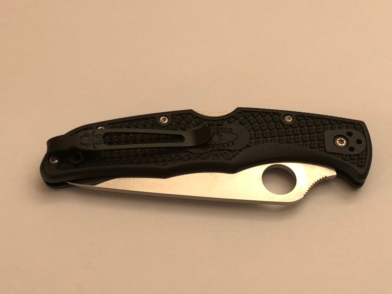 May include: A black folding knife with a silver blade and a black handle. The handle has a textured pattern and the Spyderco logo is visible on the clip.