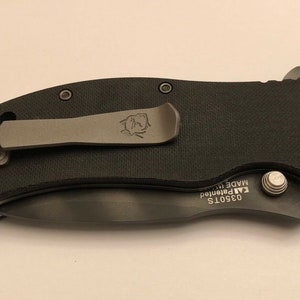 Puede incluir: Cuchillo de bolsillo negro con una pinza plateada y un tornillo plateado. El cuchillo tiene el texto "03501S KAI Patented MADE IN" en el lateral.