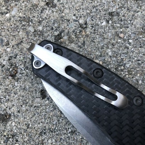 May include: A close-up of a black carbon fiber pocket knife with a silver clip. The clip is attached to the knife with two screws.