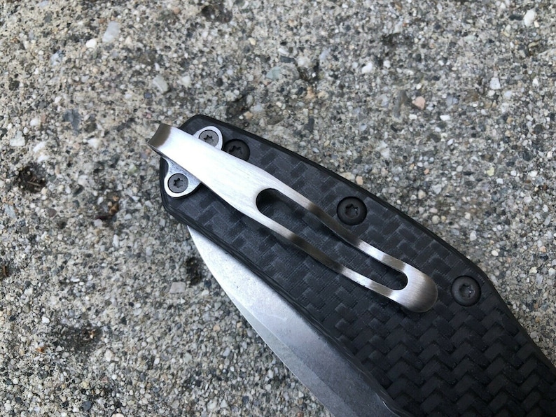 May include: A close-up of a black carbon fiber pocket knife with a silver clip. The clip is attached to the knife with two screws.