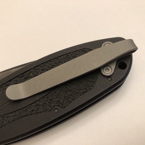 May include: A black pocket knife with a textured handle and a silver pocket clip. The knife has a black blade and a silver pocket clip.