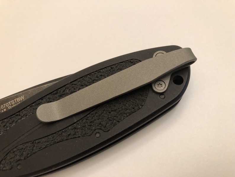 May include: A black pocket knife with a textured handle and a silver pocket clip. The knife has a black blade and a silver pocket clip.