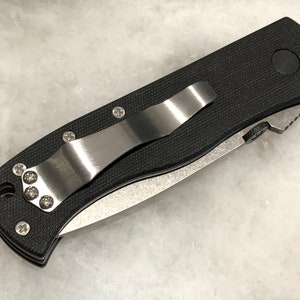 May include: A black pocket knife with a stainless steel clip and a black handle. The knife has a partially serrated blade.