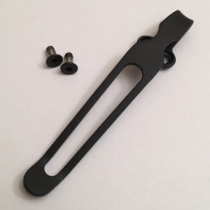 May include: Black metal pocket clip with two screws. The clip has a rounded end and a long, narrow opening.