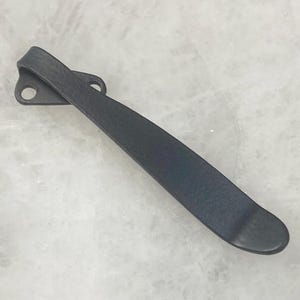May include: A matte black pocket knife clip with two mounting holes. The clip is curved and has a smooth, rounded end. The metal appears to have a textured finish. The clip is designed to attach to a pocket knife.